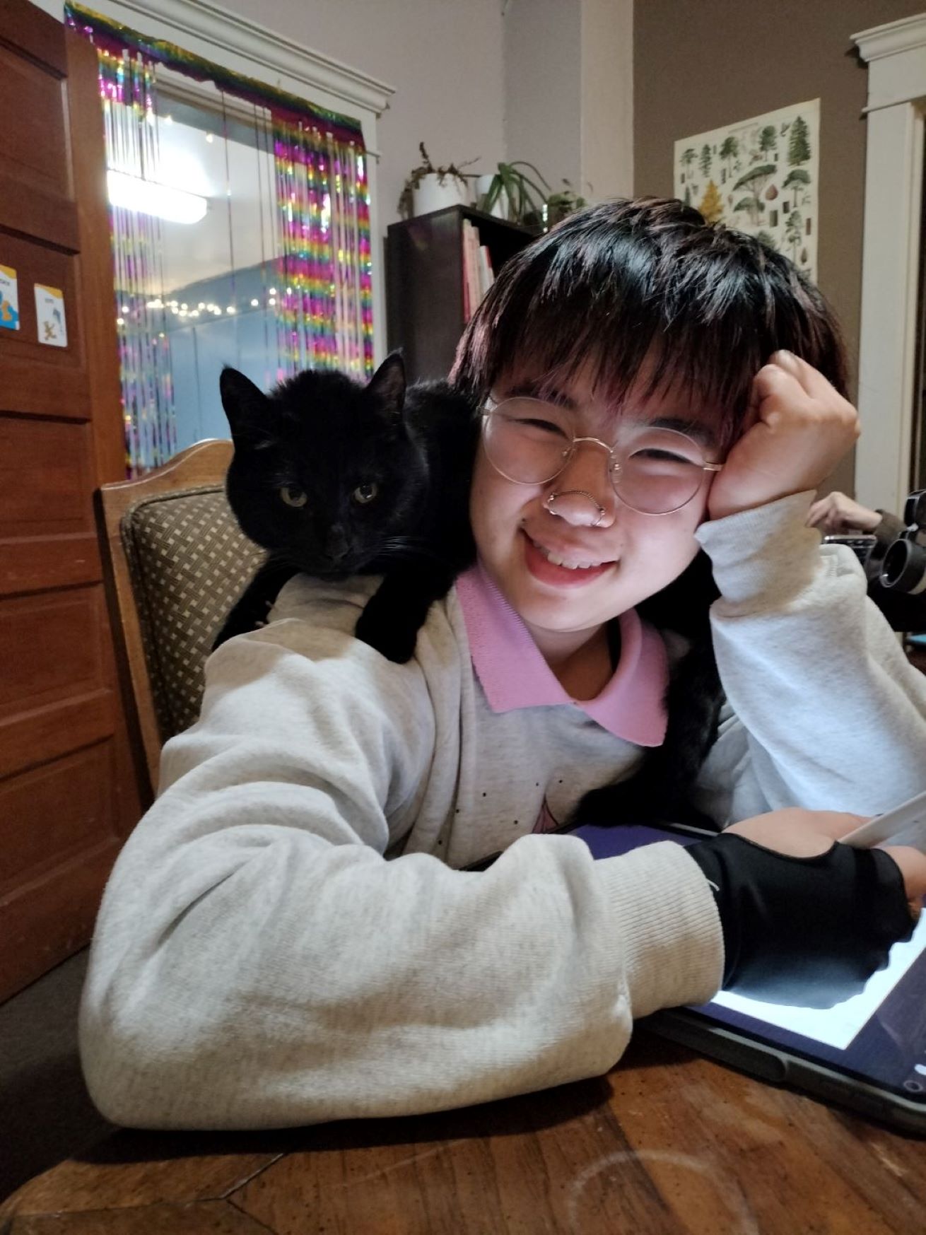 Alex wearing glasses and a white sweatshirt, smiling with black cat on right shoulder and head rested on left hand