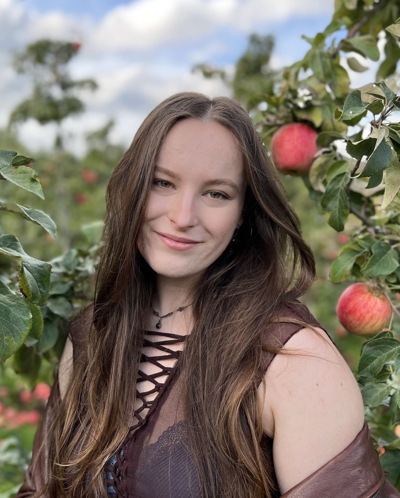 Elizabeth smiling with dark hair down and wearing a brown shirt, standing in front of an apple tree