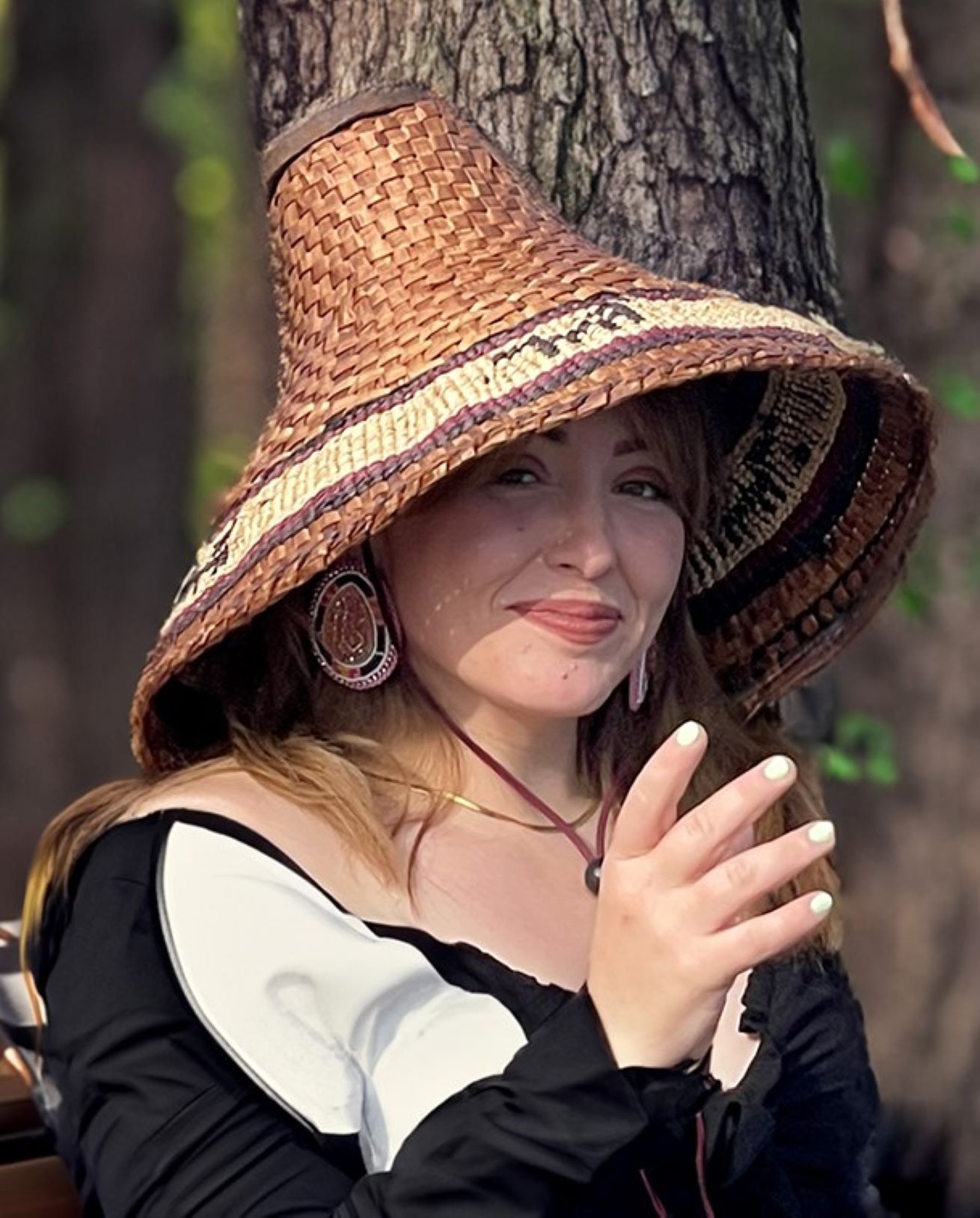 Kimy smiling, wearing a large woven brown hat and right hand lifted, standing in front of tree