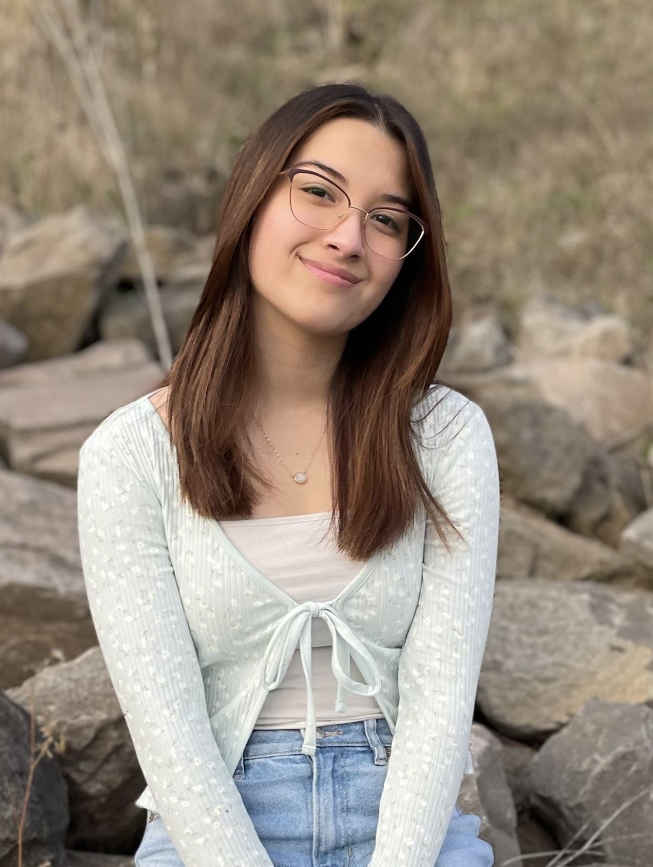 Lily smiling with glasses on, wearing a white sweater and light wash denim jeans, sitting ourside on rocks