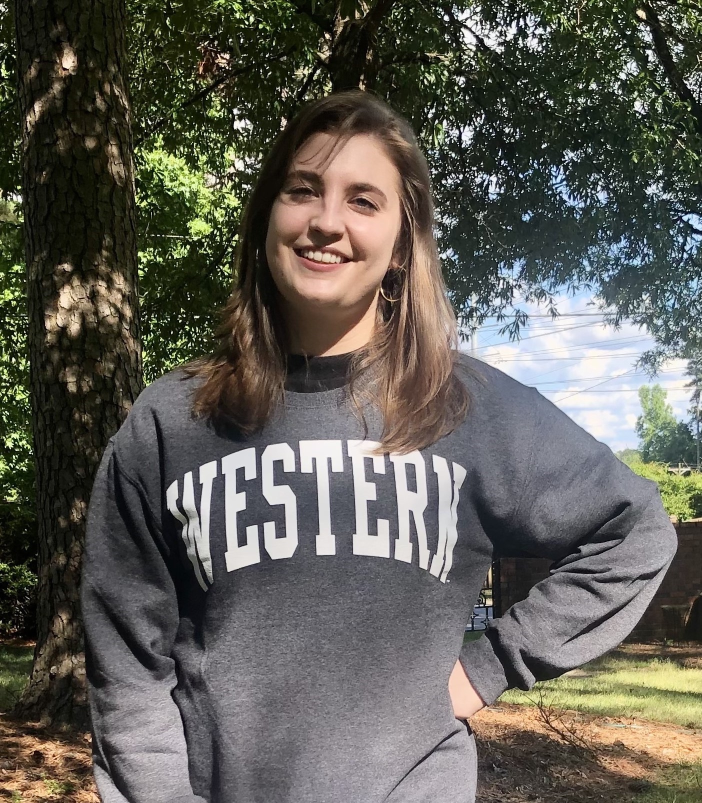 Lydia smiling and standing in front of trees wearing a grey sweatshirt with white text: "Western" and left hand on waist