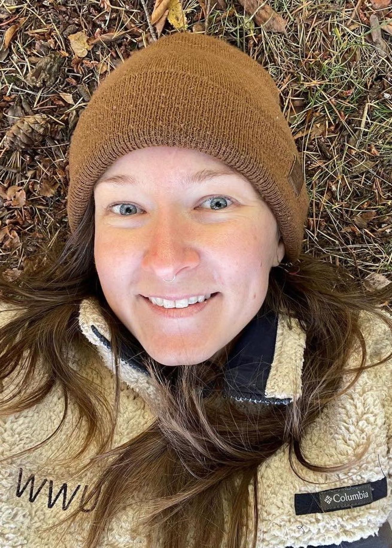 Casey smiling wearing a brown beanie and beige "WWU" pullover, laying on a pile of brown leaves and pine needles