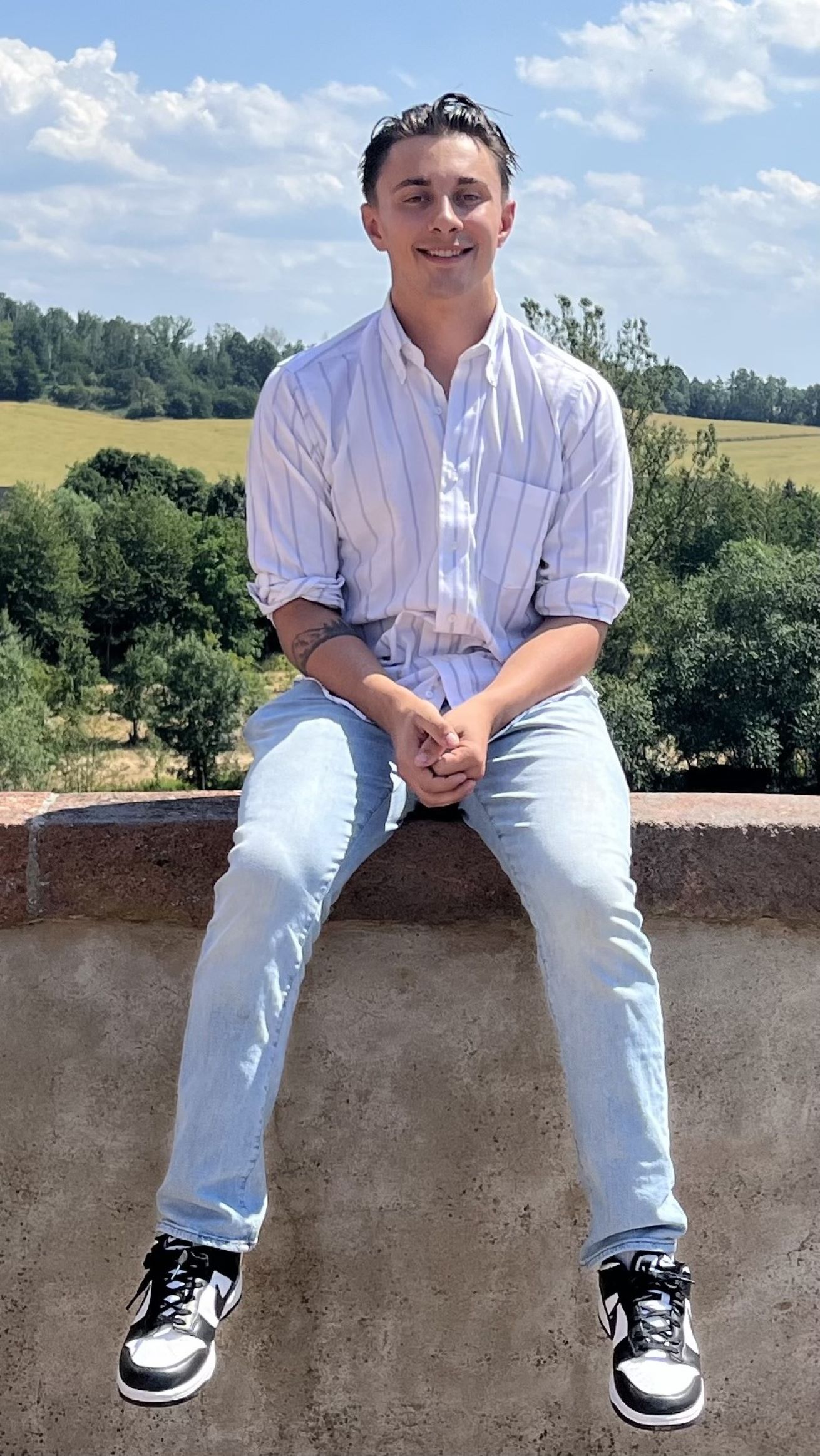 Devan Martin smiling, wearing a white shirt and light wash jeans, sitting on a ledge in front of trees in the background