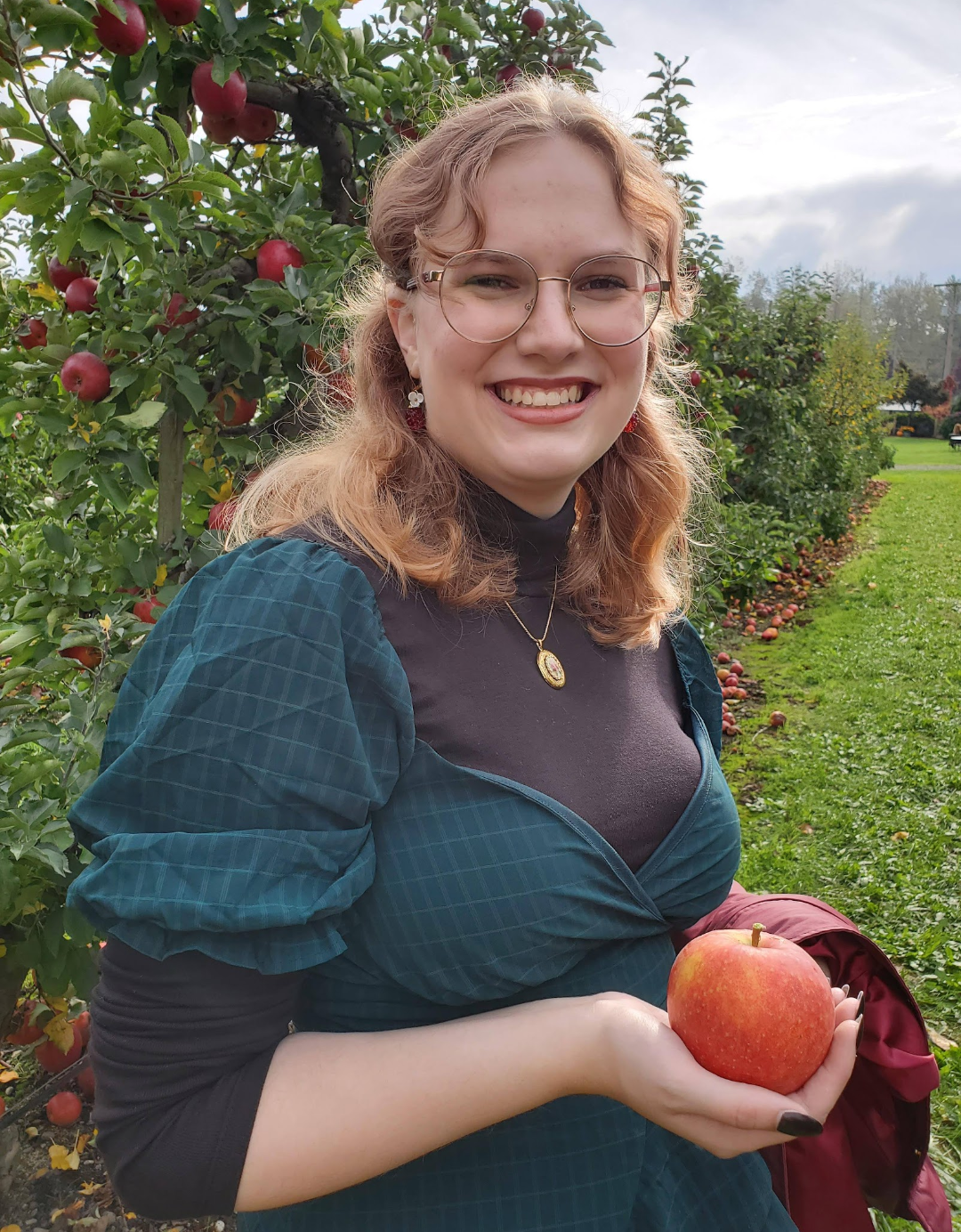 Audrey smiling wearing glasses and a blue/green dress and purple shirt, holding an apple