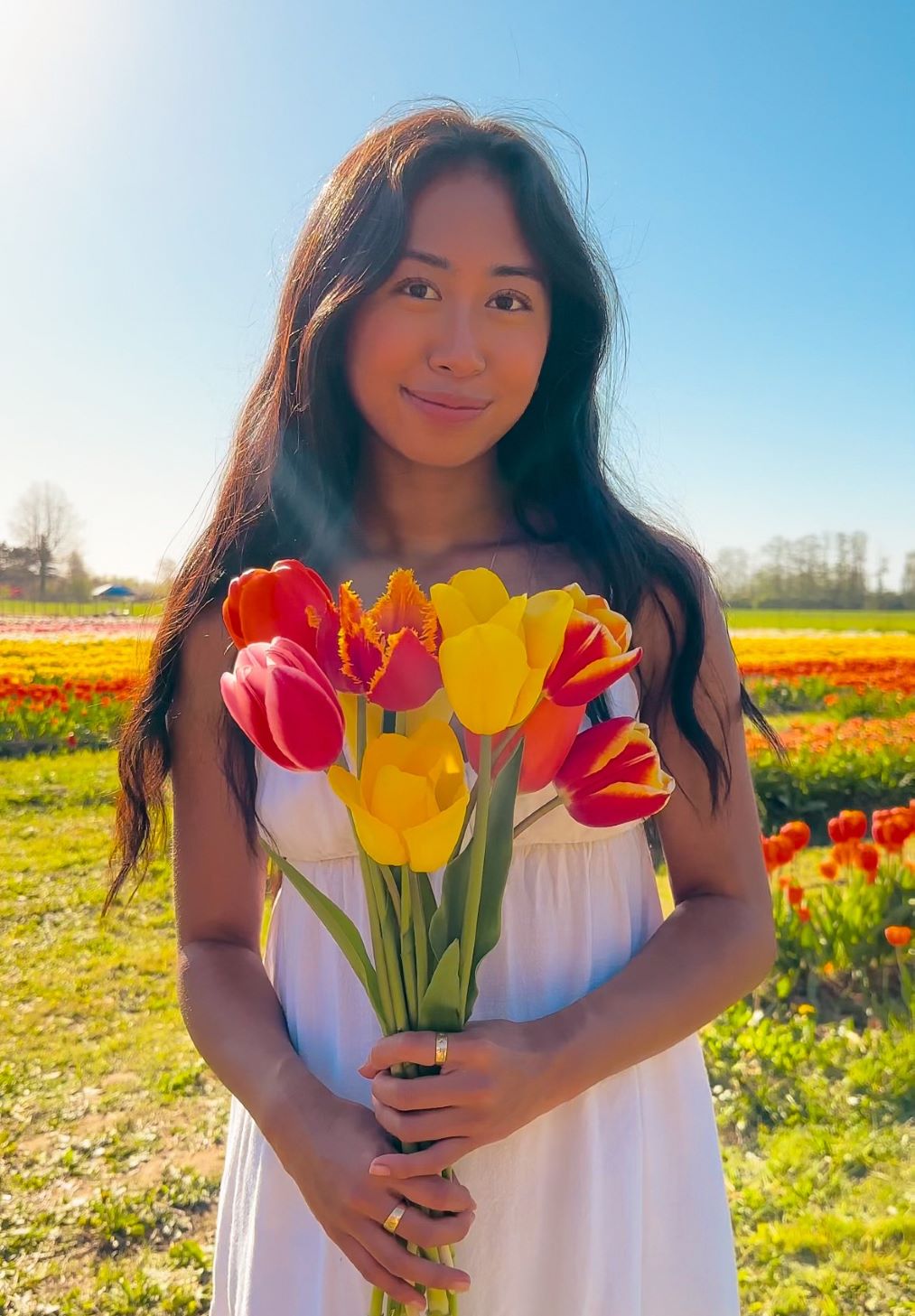 Malia wearing a white dress, smiling and standing in front of blue sky and tulip fields, holding a bouquet of pink, red, and yellow tulips 