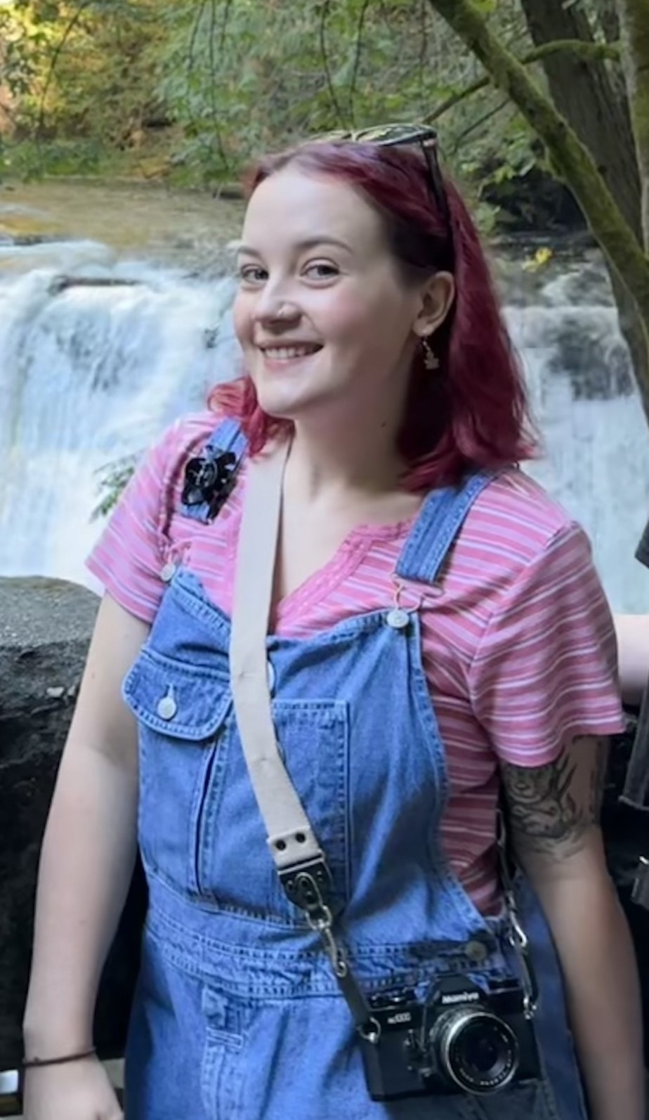 Abi smiling, wearing a striped pink shirt and denim overalls and a camera around neck; standing in front of waterfall